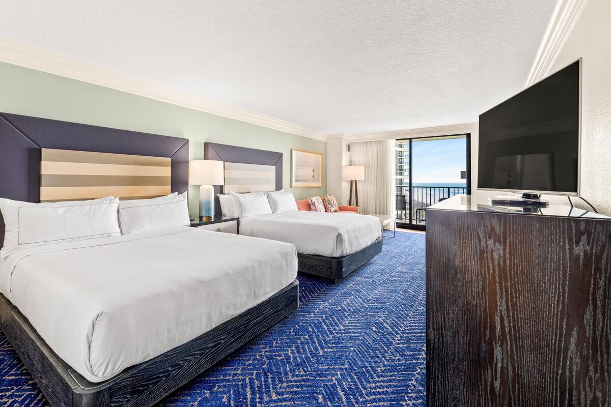 A hotel room with two beds, a desk, a TV, and a balcony with a view.