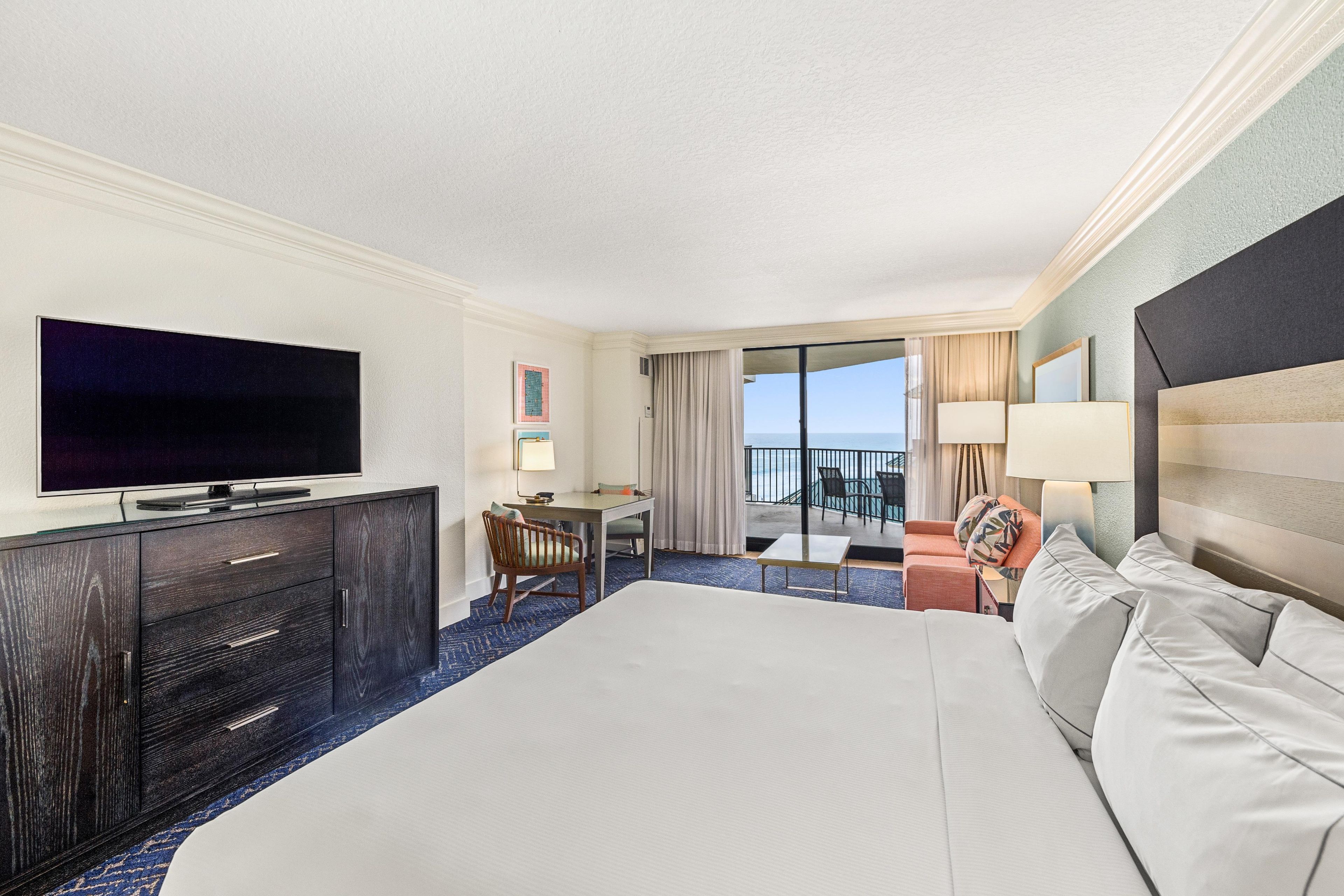 A hotel room with a large bed, a flat-screen TV, a desk with a chair, a small table, and a balcony with a view of the ocean.