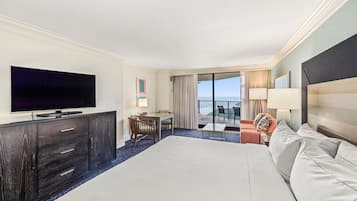 A hotel room with a large bed, a flat-screen TV, a desk with a chair, a small table, and a balcony with a view of the ocean.