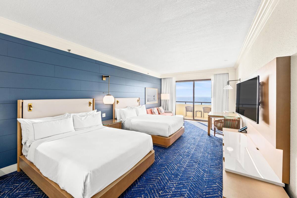 A hotel room with two beds, a TV, a desk, and a balcony with a view.