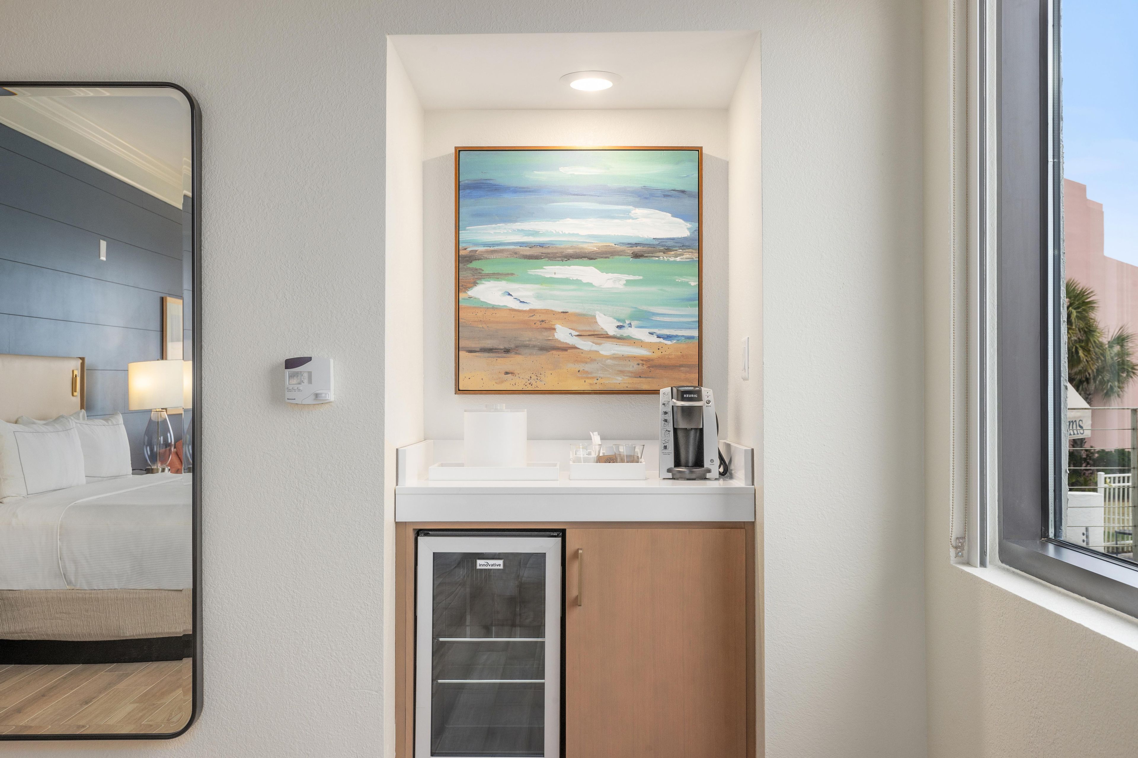 Une chambre d’hôtel avec un lit, une machine à café, un tableau représentant une scène de plage et une fenêtre donnant sur des palmiers.