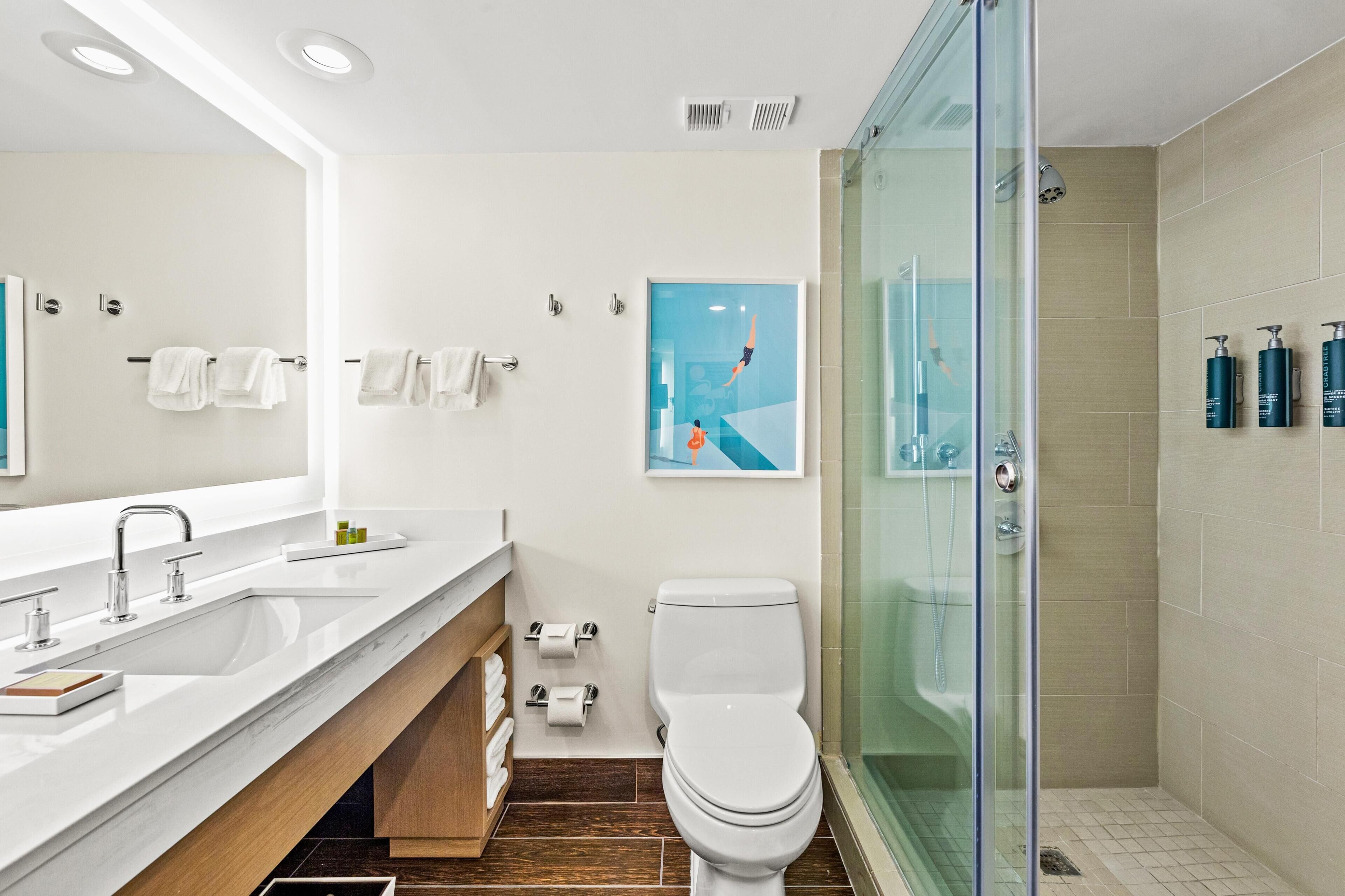A modern bathroom with a glass shower enclosure, a white sink with a wooden vanity, and a toilet with a roll of toilet paper beside it.