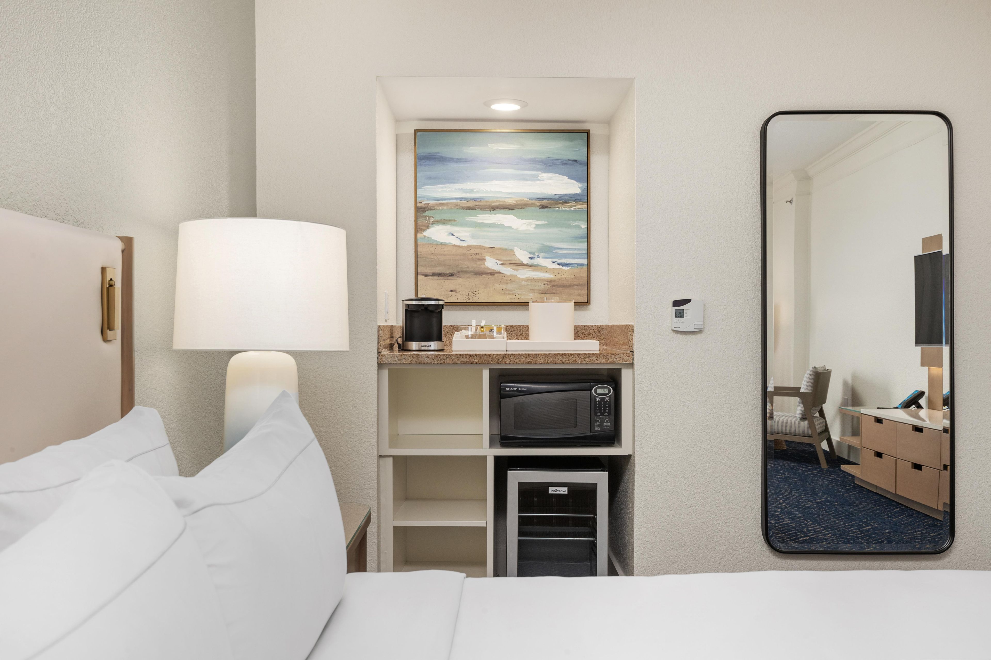 A hotel room with a bed, a painting of a beach, a microwave, and a mirror.