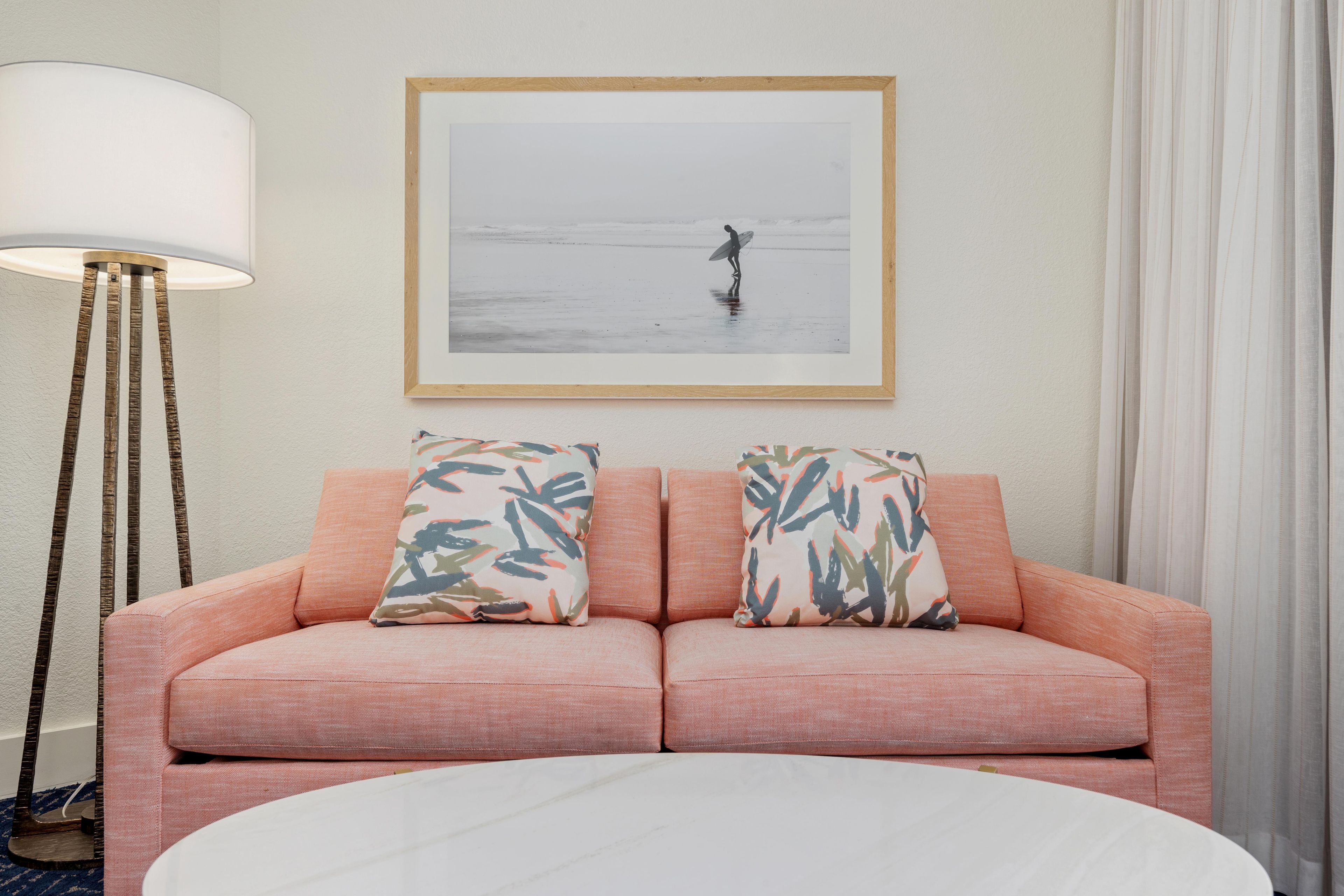 A room with a pink sofa, two patterned pillows, a framed picture of a beach scene, a floor lamp, and a round white table.