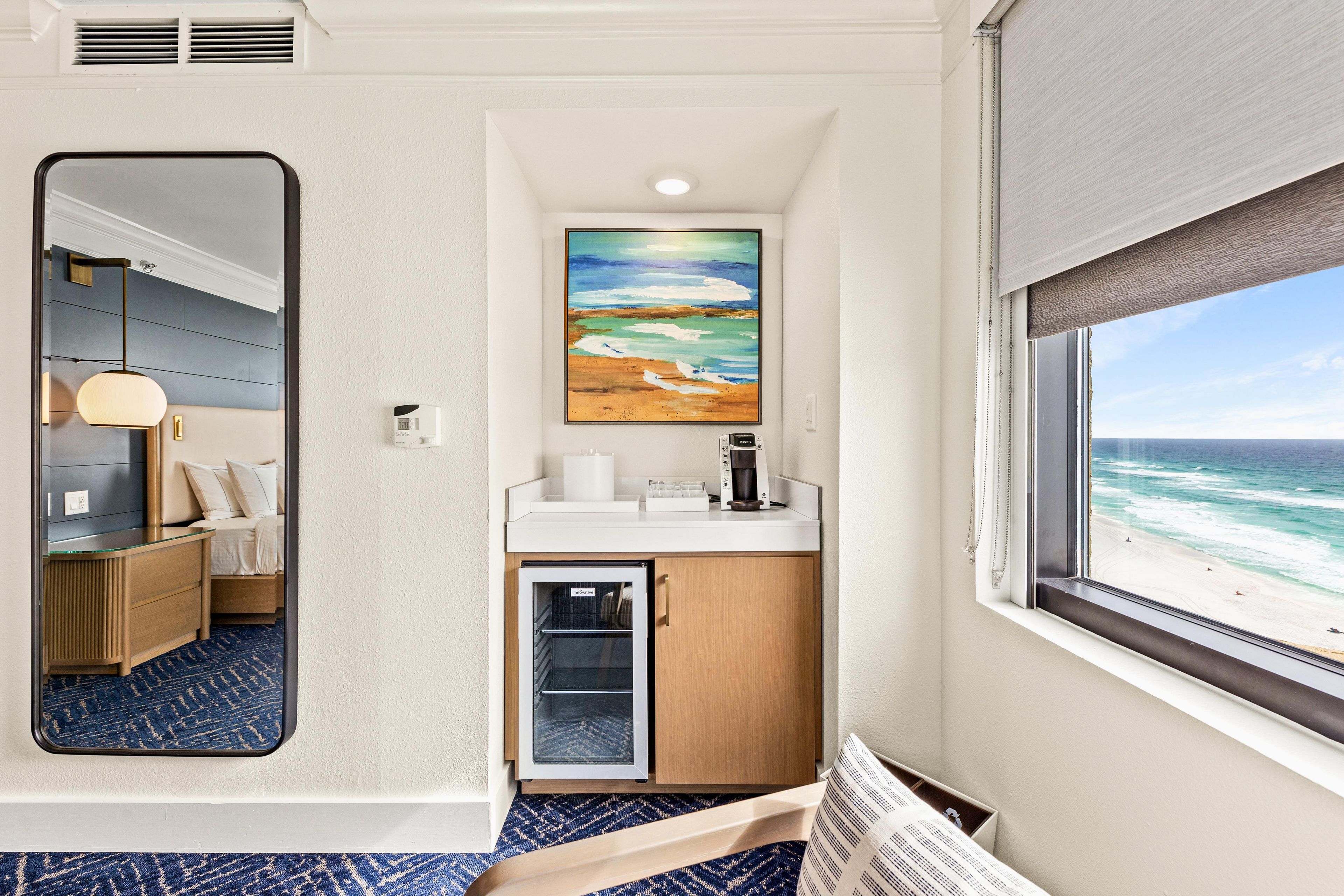 A hotel room with a large mirror, a painting of a beach, a coffee machine, and a view of the ocean.