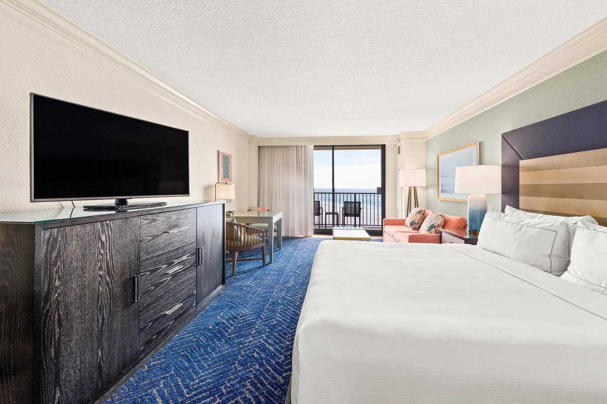 A hotel room with a large bed, a flat-screen TV, a desk with a chair, a balcony with a view, and a large window.