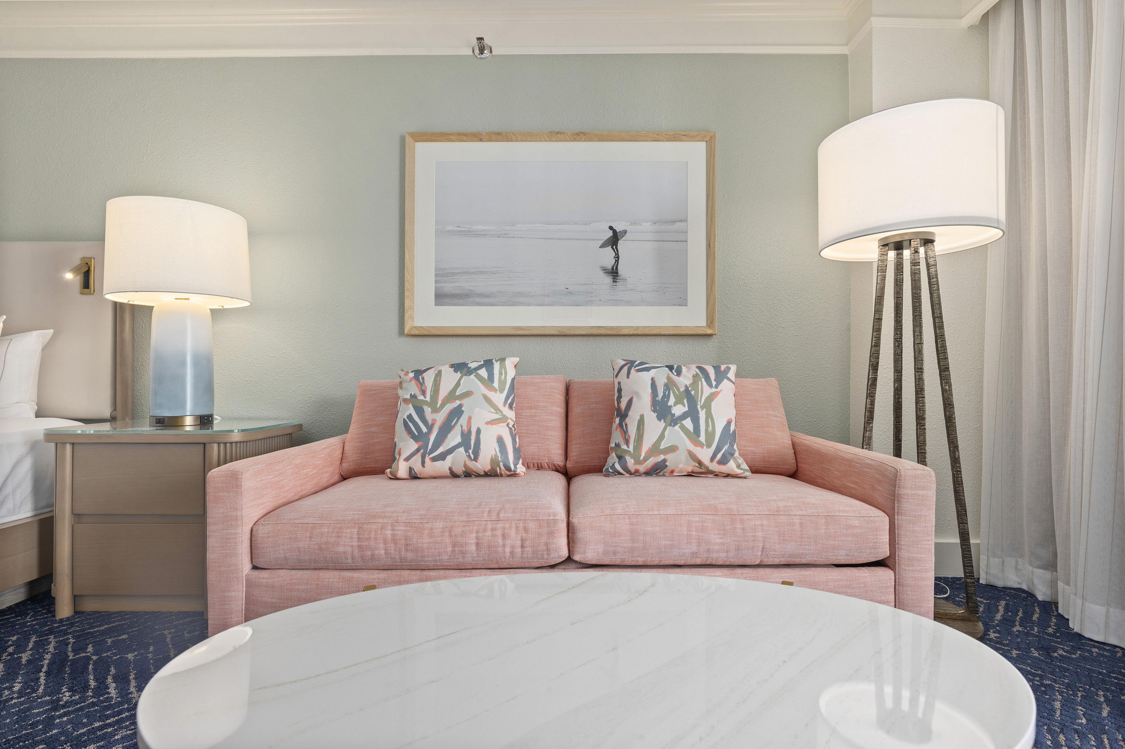 Habitación de hotel con sofá rosa, dos almohadas estampadas, un cuadro enmarcado de un surfista, una mesa redonda blanca y dos lámparas de pie.