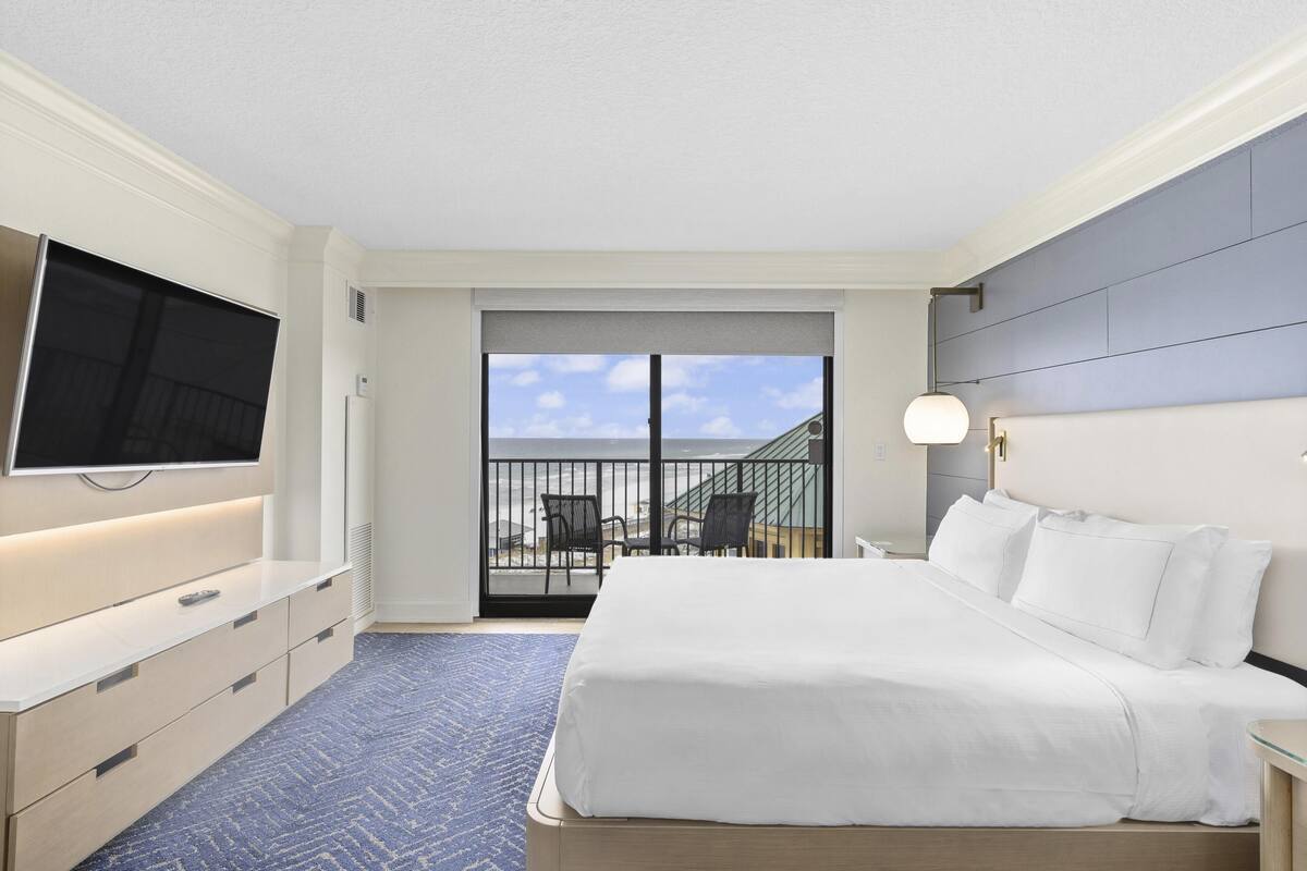A hotel room with a large bed, a TV mounted on the wall, a balcony with chairs, and a view of the sky.