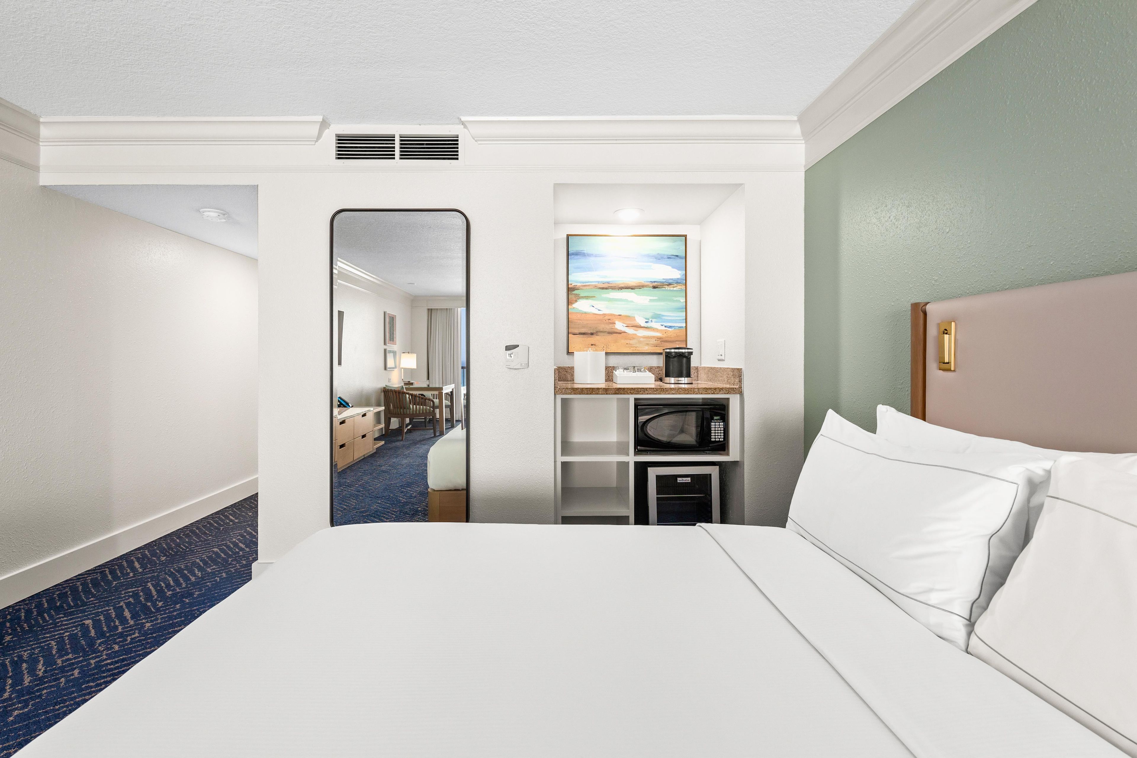 Quarto de hotel com uma cama, escrivaninha com micro-ondas, pintura e vista para um corredor.
