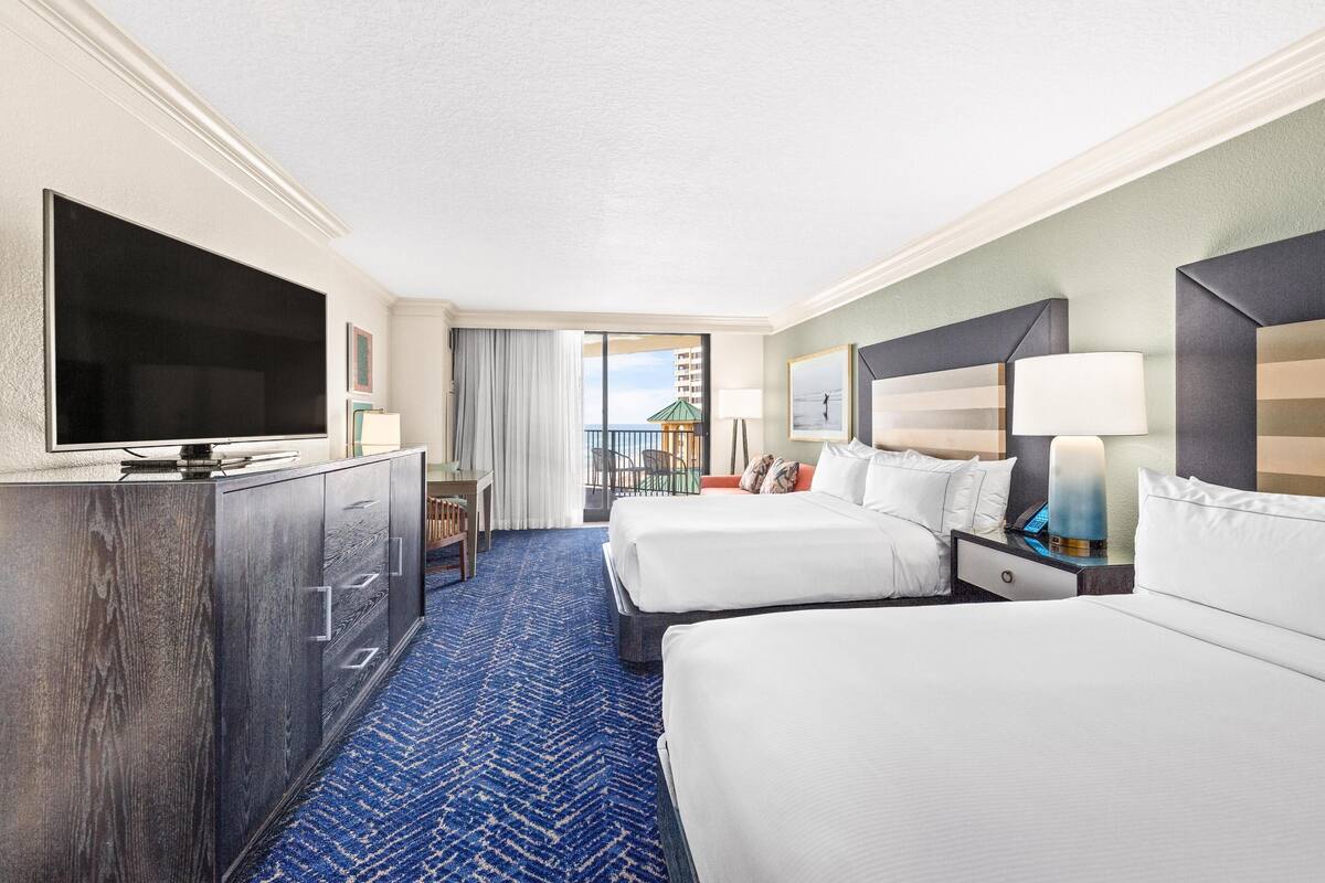 A hotel room with a large flat-screen TV, two beds, a desk, and a balcony with a view of the sea.