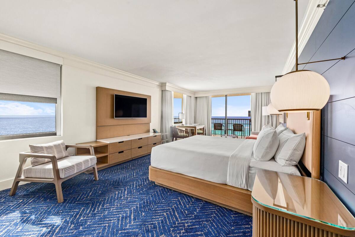 A modern hotel room with a large bed, a TV, a desk, and a view of the ocean.