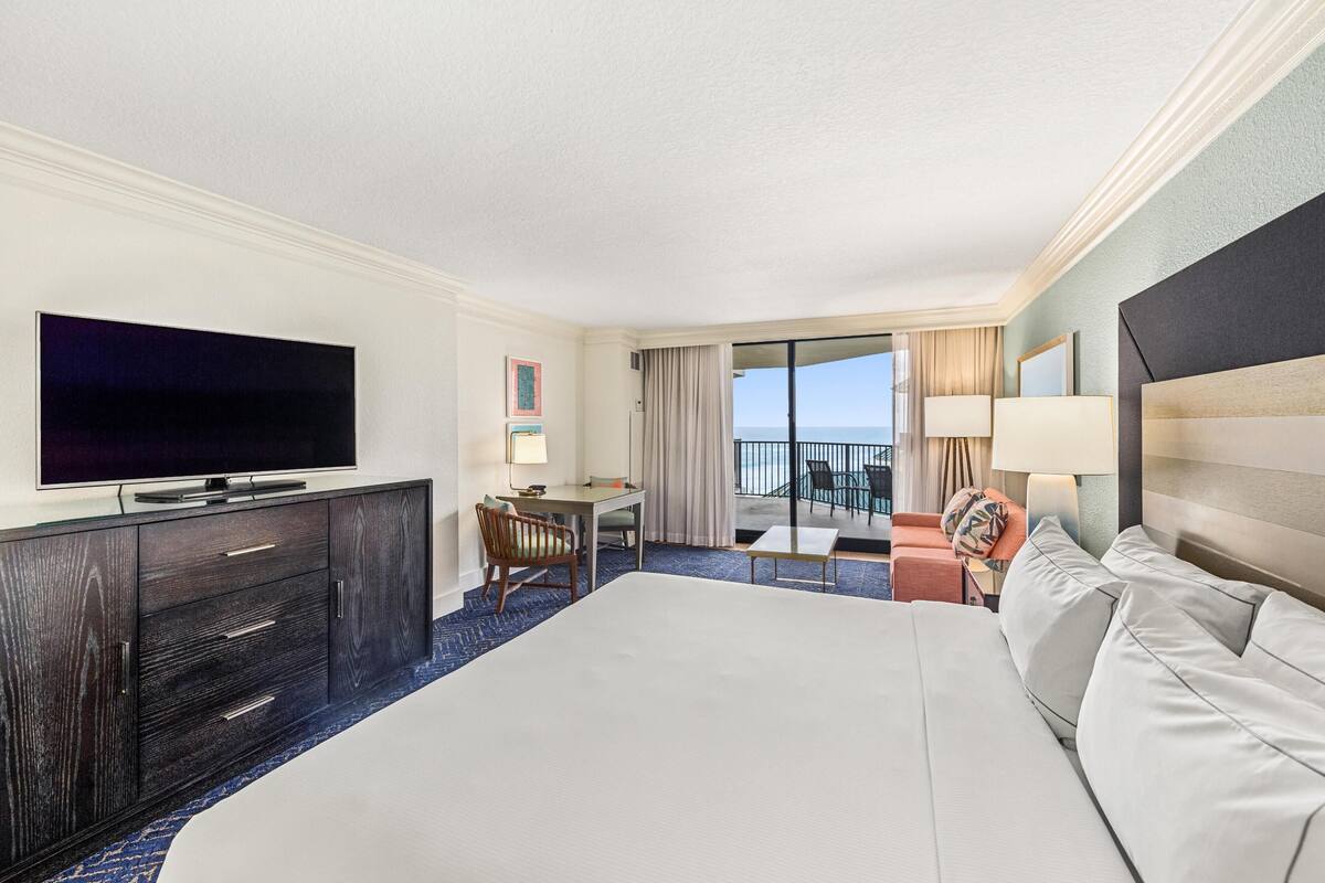 A hotel room with a large bed, a flat-screen TV, a desk with a chair, a balcony with a view of the ocean, and a large window.