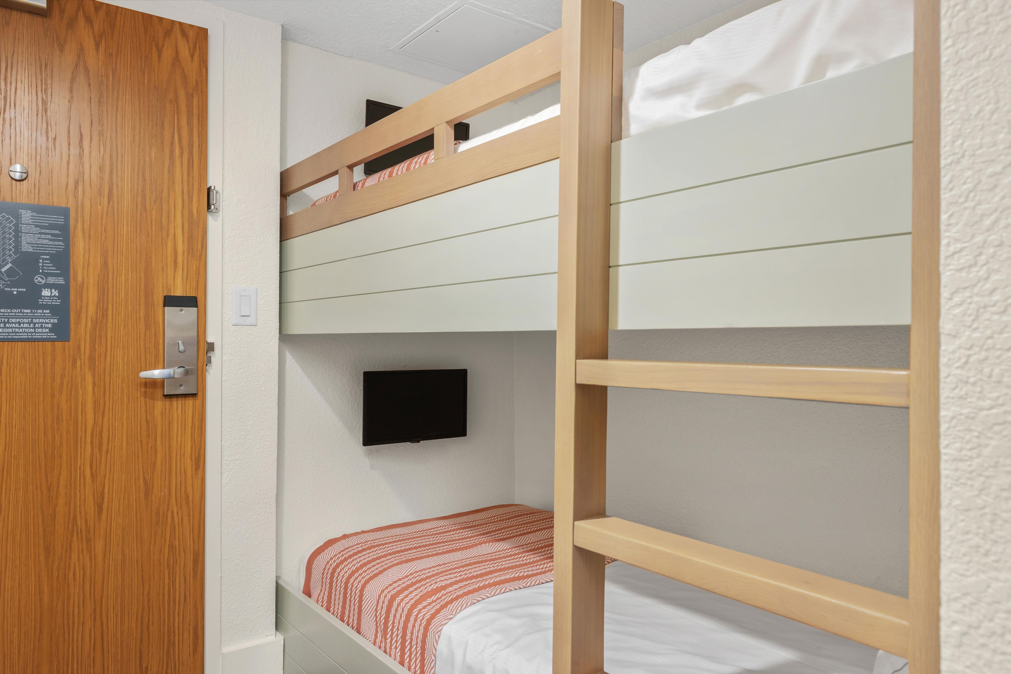 Habitación con literas, una escalera de madera, un televisor en la pared y una puerta con un letrero.