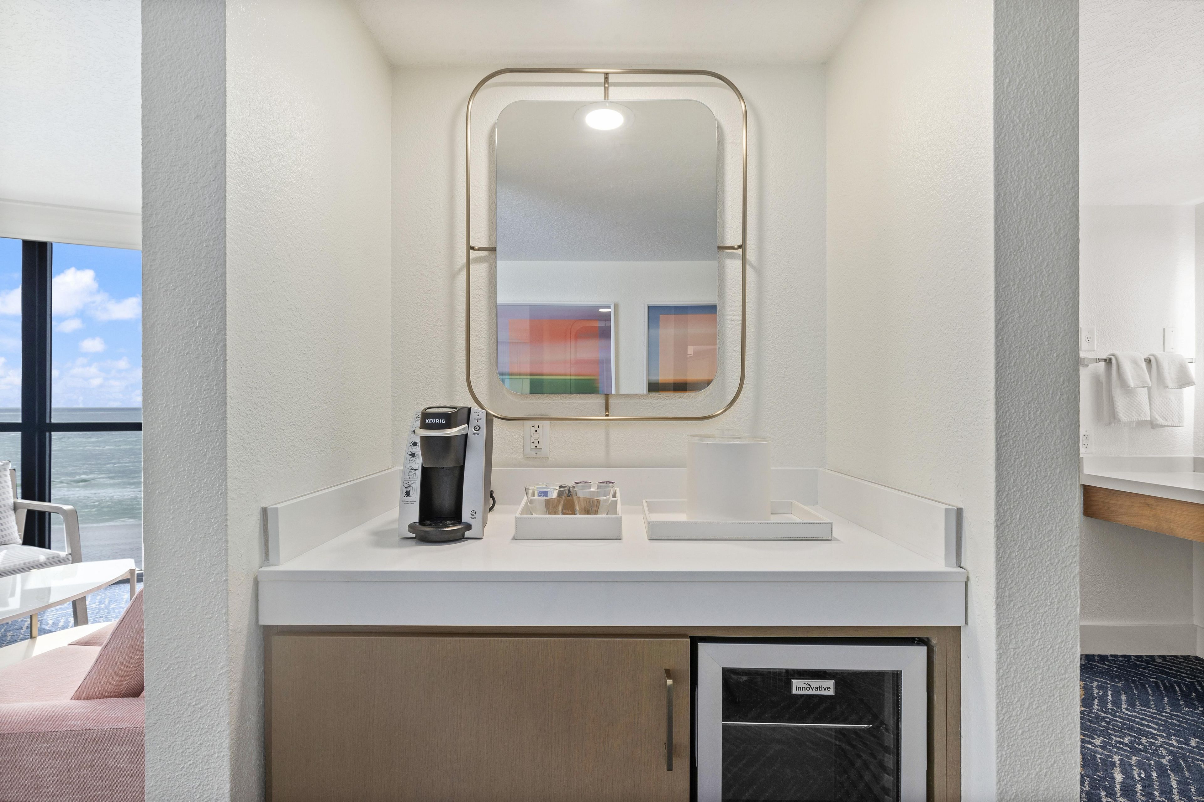 Quarto de hotel com minibar branco, cafeteira, espelho e vista para o oceano.