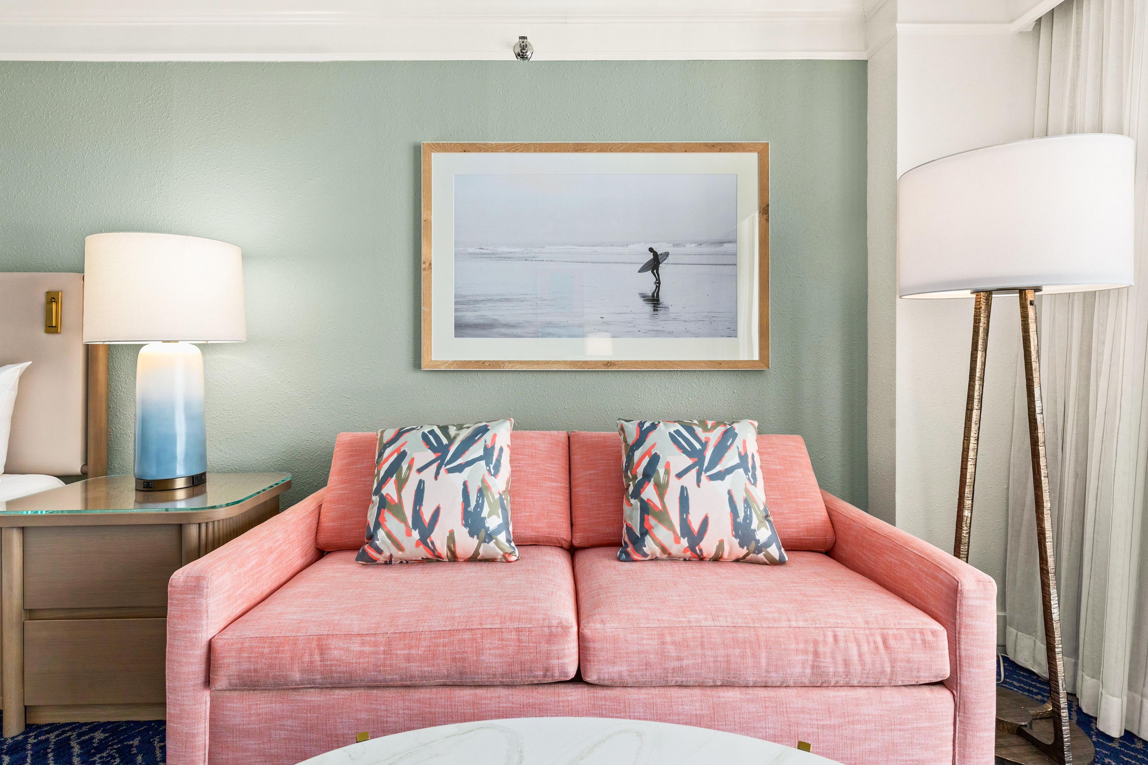 Une chambre d’hôtel avec un canapé rose, deux coussins à motifs, une photographie encadrée accrochée au mur, une lampe de chevet et une grande lampe sur pied.