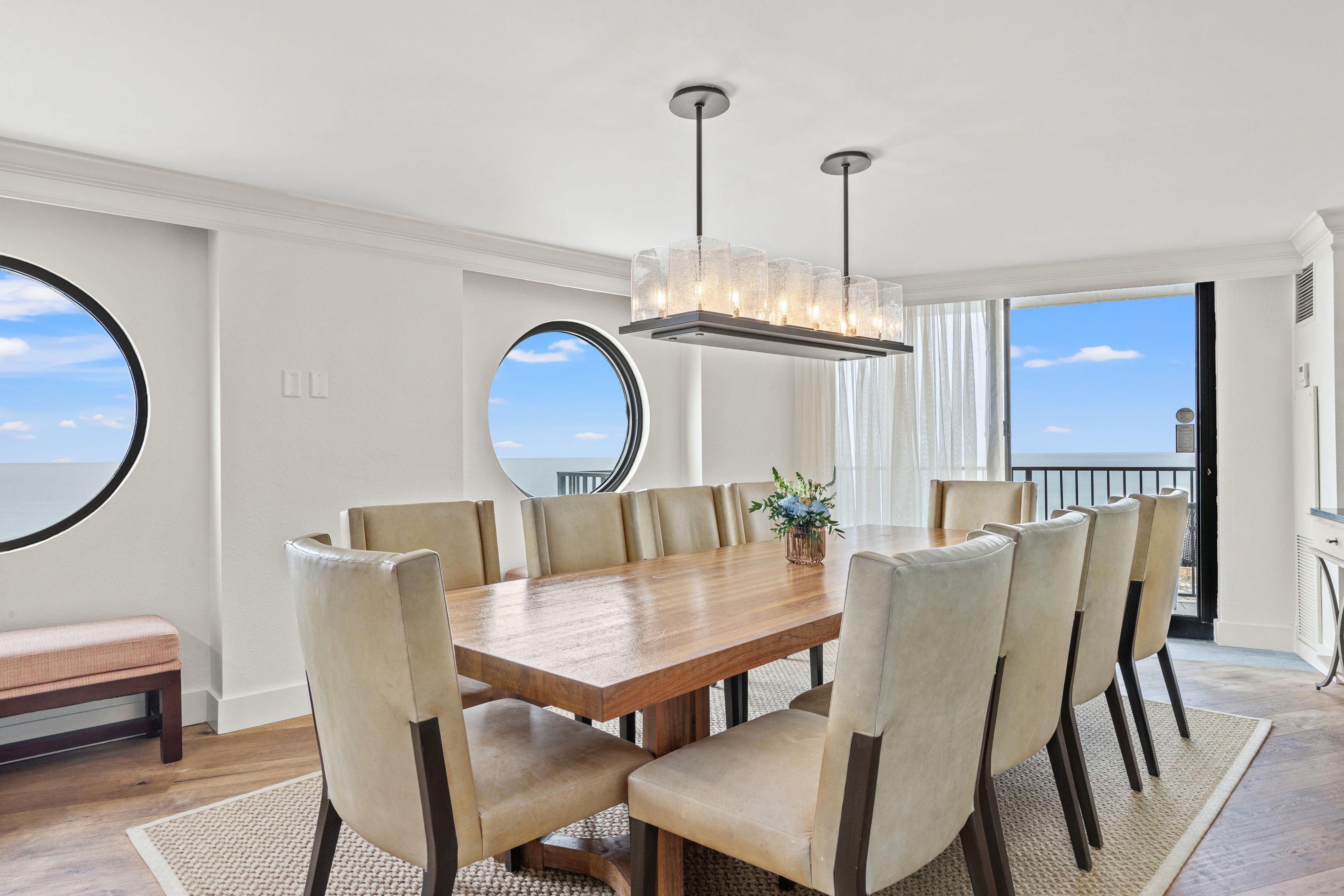 Un coin repas avec une table et des chaises en bois, un lustre et une vue sur la mer grâce à de grandes fenêtres.