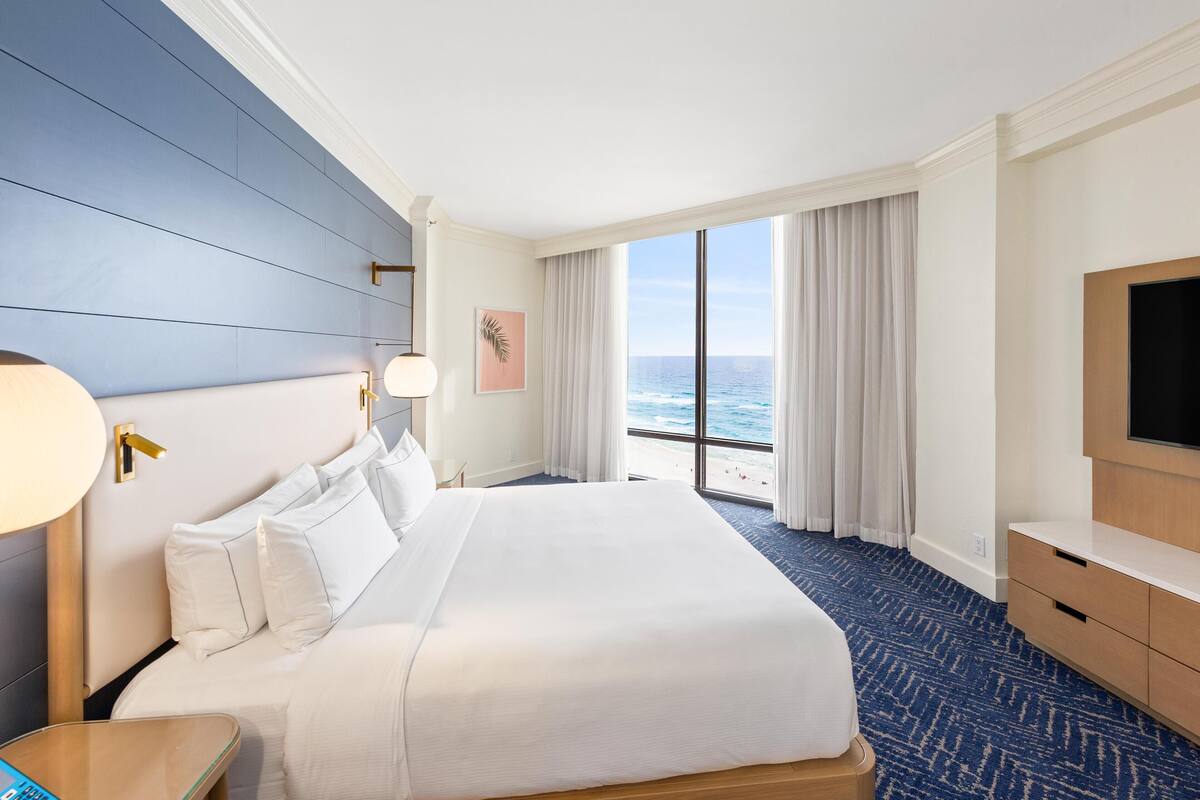 A hotel room with a large bed, a TV, a desk, and a view of the ocean.