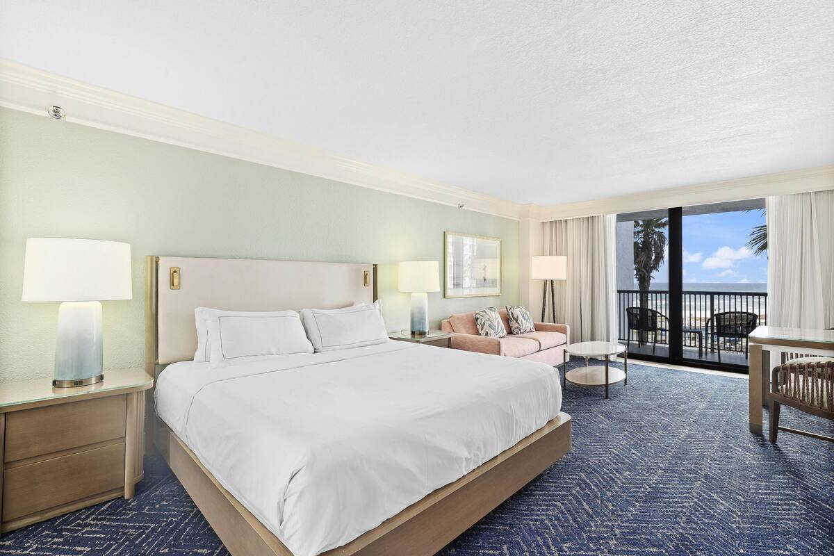A hotel room with a large bed, a sofa, a small table, and a balcony with a view.