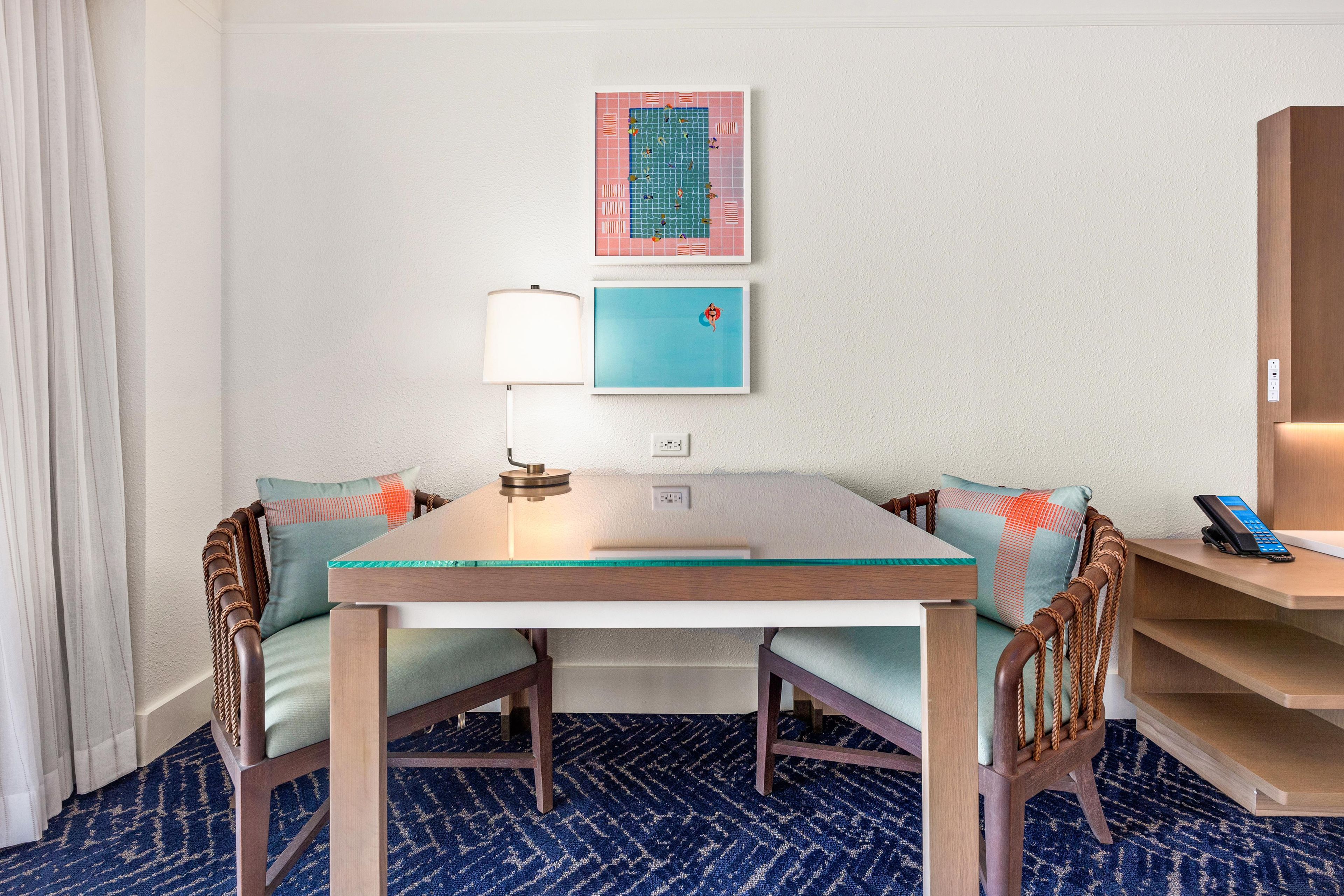A hotel room with a dining table, chairs, a lamp, a phone, and artwork on the wall.