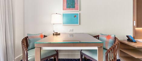 A hotel room with a dining table, chairs, a lamp, a phone, and artwork on the wall.