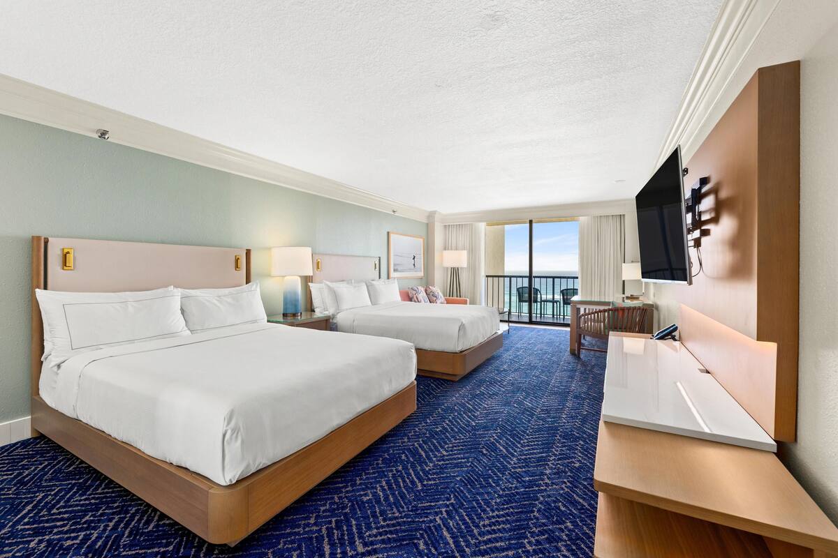 A hotel room with two beds, a desk, a TV, and a balcony with a view.