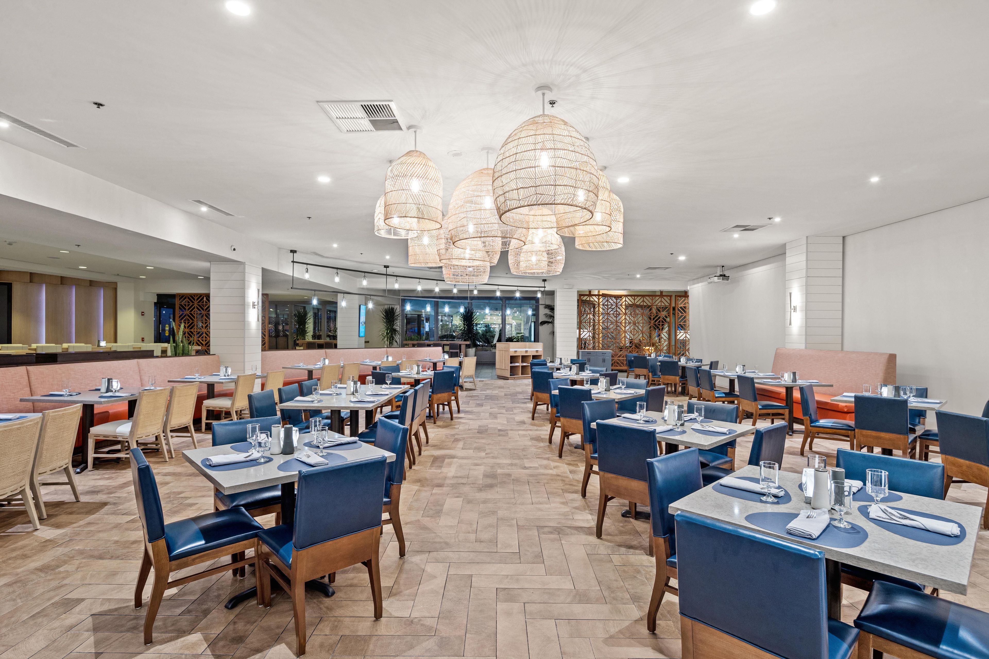 A spacious restaurant with multiple dining tables and chairs, large hanging light fixtures, and a view of the cityscape through large windows.
