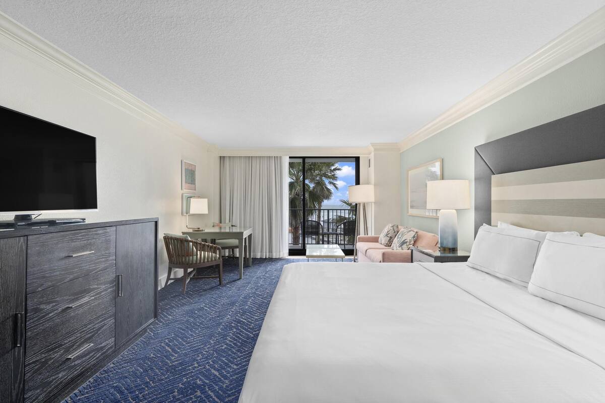 A hotel room with a large bed, a desk, a chair, a television, and a balcony with a view of palm trees.