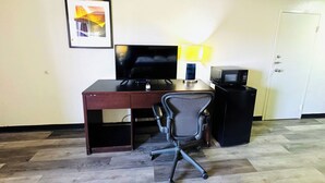 A hotel room with a wooden desk, a black office chair, a flat-screen TV, a lamp, a microwave, and a door. - Newnan Lodge (Newnan)