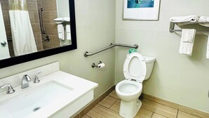 A bathroom with a white sink, a toilet, a shower with a glass door, a handrail, and a framed picture on the wall. - Newnan Lodge (Newnan)