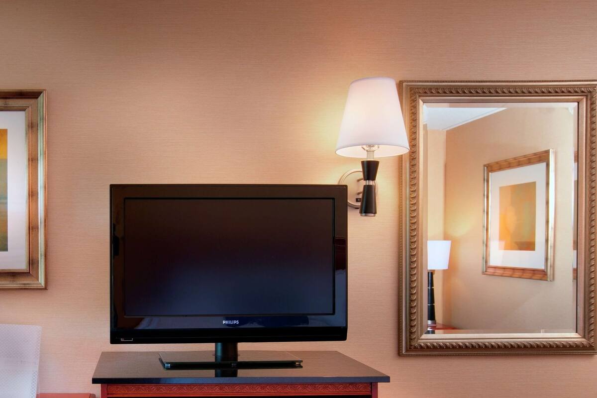A hotel room with a flat-screen TV, a framed abstract painting, a wall-mounted lamp, and a mirror.