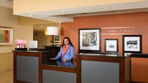 A hotel reception desk with a receptionist, framed pictures on the wall, and a 'Welcome to Alexandria' sign.