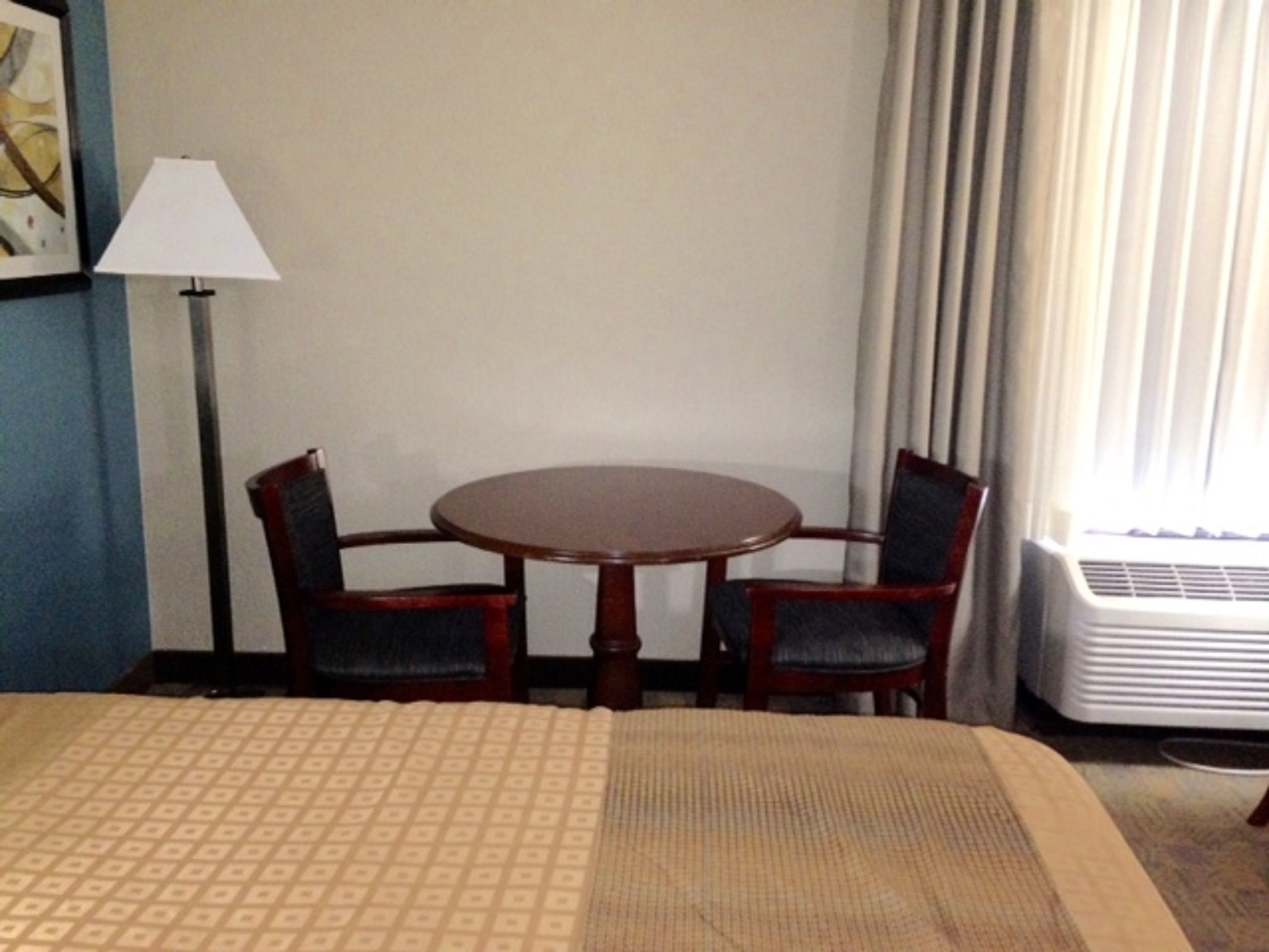 Habitación de hotel con cama, una mesa redonda con dos sillas, una lámpara y un ventanal con cortinas.