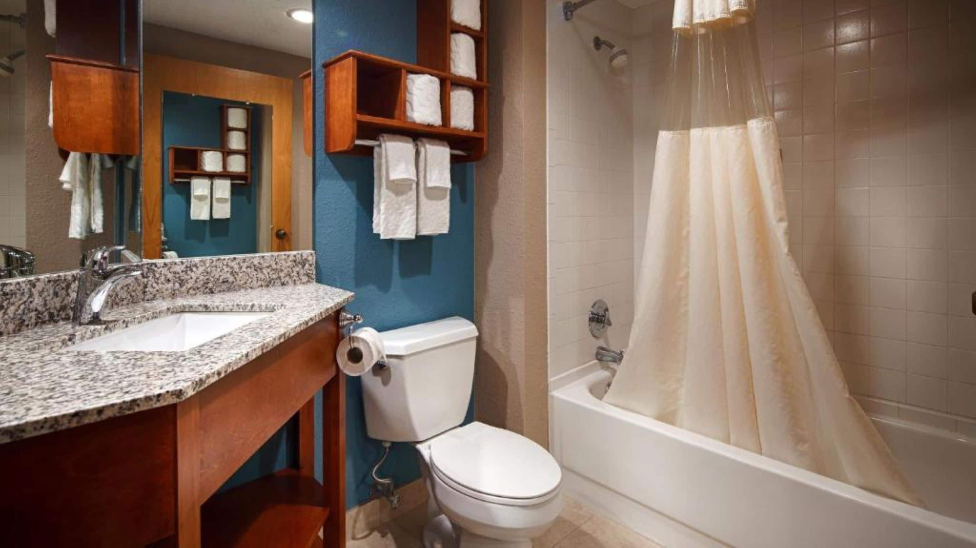 A bathroom with a granite countertop sink, a toilet, a shower with a beige curtain, and a wooden medicine cabinet with towels.