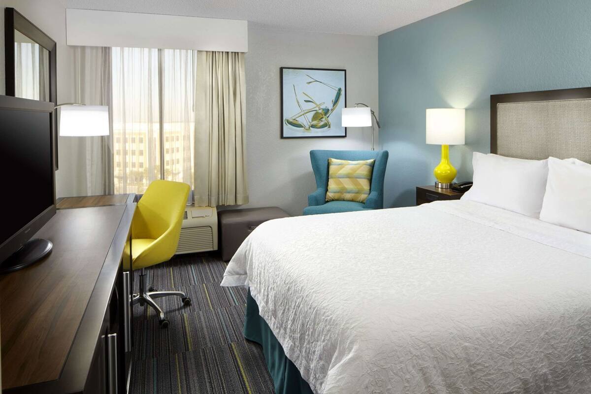 A hotel room with a bed, a desk with a chair, a television, a lamp, and a painting on the wall.