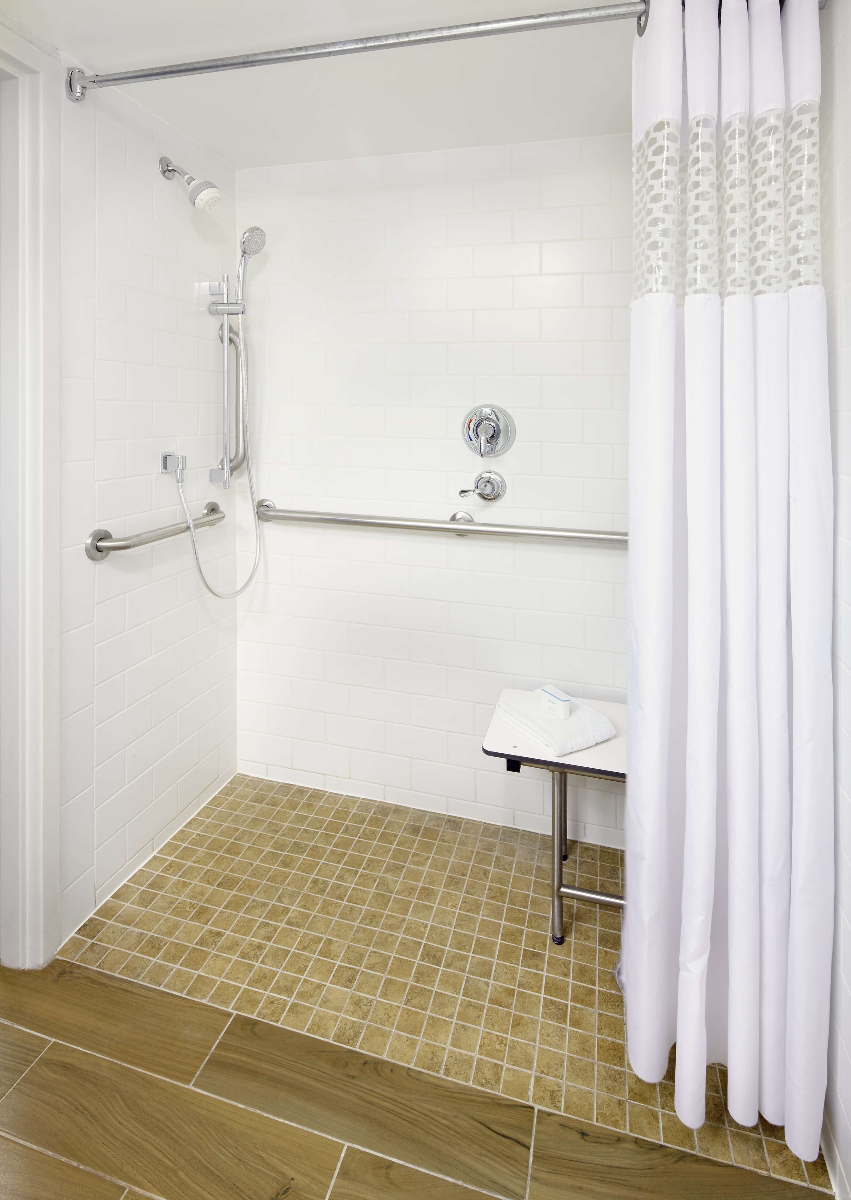 Un baño con ducha, barra de apoyo y lavamanos pequeño con toalla.