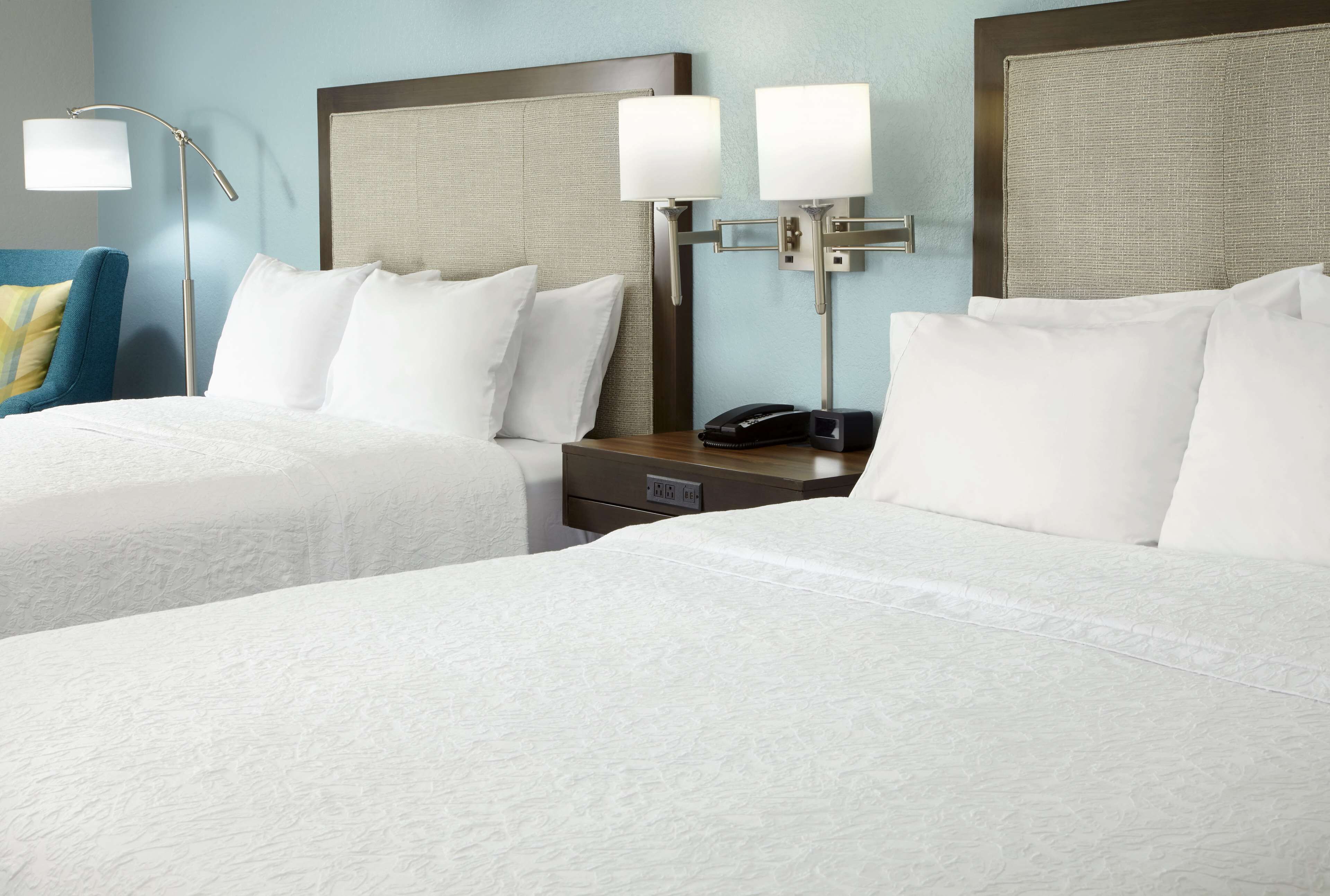 A hotel room with two beds, a nightstand with a phone, and a wall-mounted lamp.
