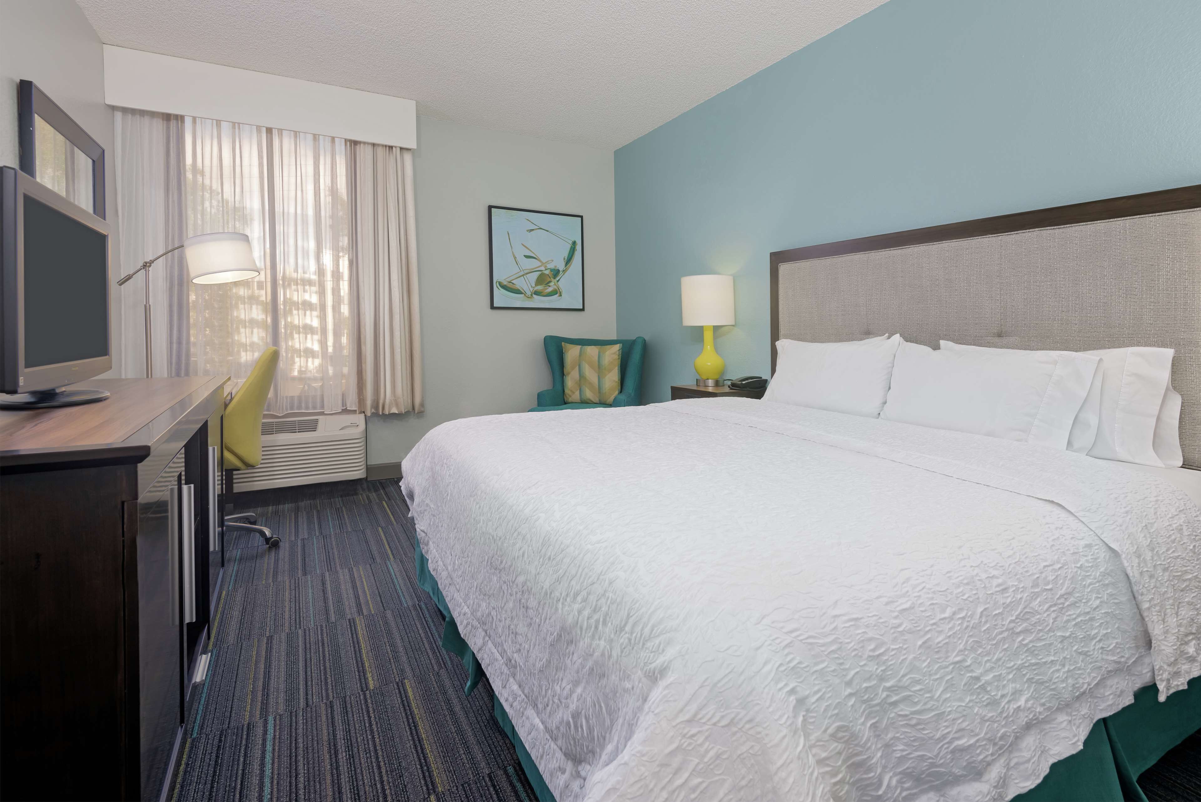 A hotel room with a large bed, a desk with a computer, a chair, a lamp, and a window with curtains.