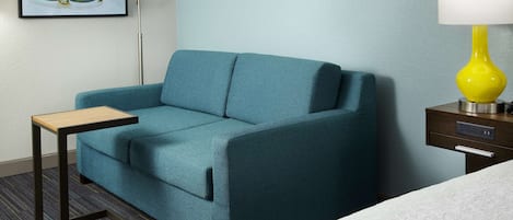 A teal sofa, a small wooden table, a yellow lamp, and a white bed in a hotel room.