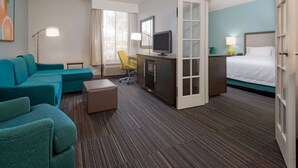 Una habitación de hotel con un sofá color turquesa, un escritorio con una computadora, una cama y un ventanal con cortinas.
