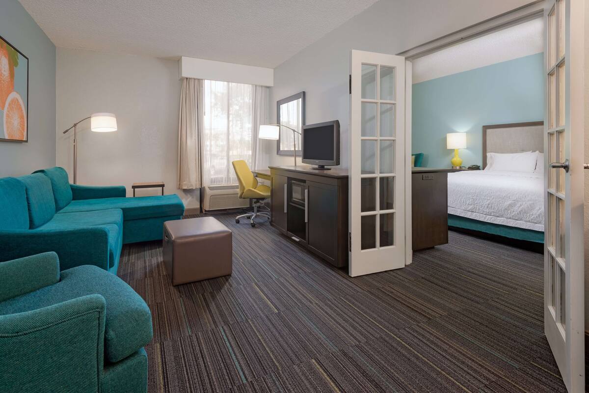 Una habitación de hotel con un sofá color turquesa, un escritorio con una computadora, una cama y un ventanal con cortinas.