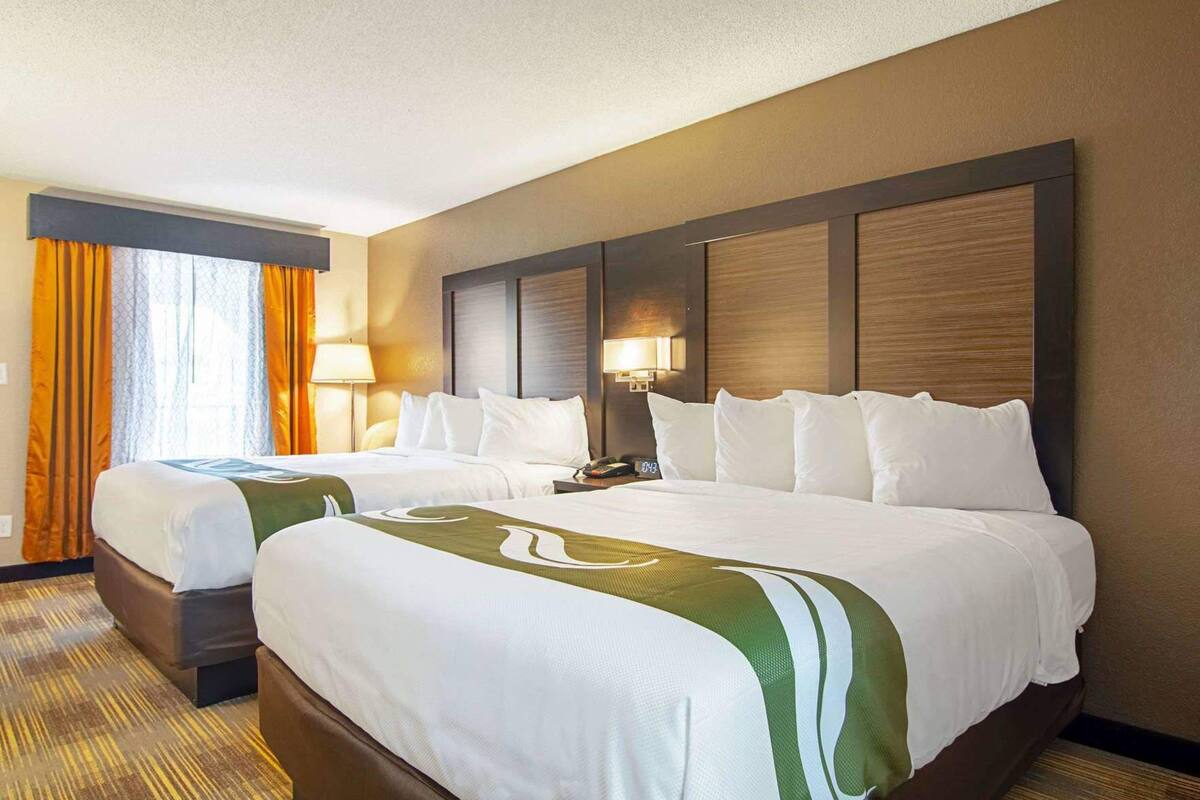 A hotel room with two beds, a large window with curtains, a nightstand, and a wall-mounted lamp.