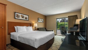 Habitación de hotel con una cama grande, un escritorio con televisor, una silla y un ventanal con cortinas.