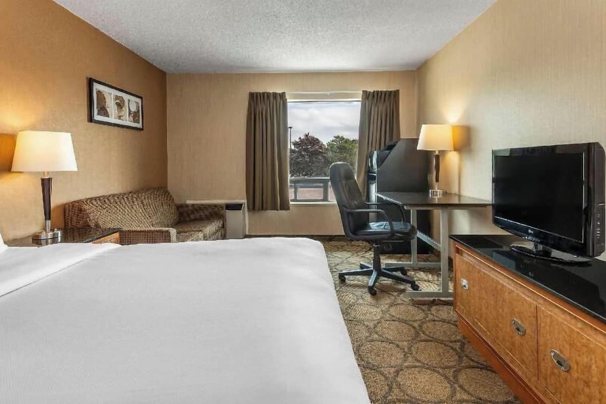 Une chambre d’hôtel avec un lit, un bureau avec une télévision, une chaise, une lampe et une fenêtre avec des rideaux.