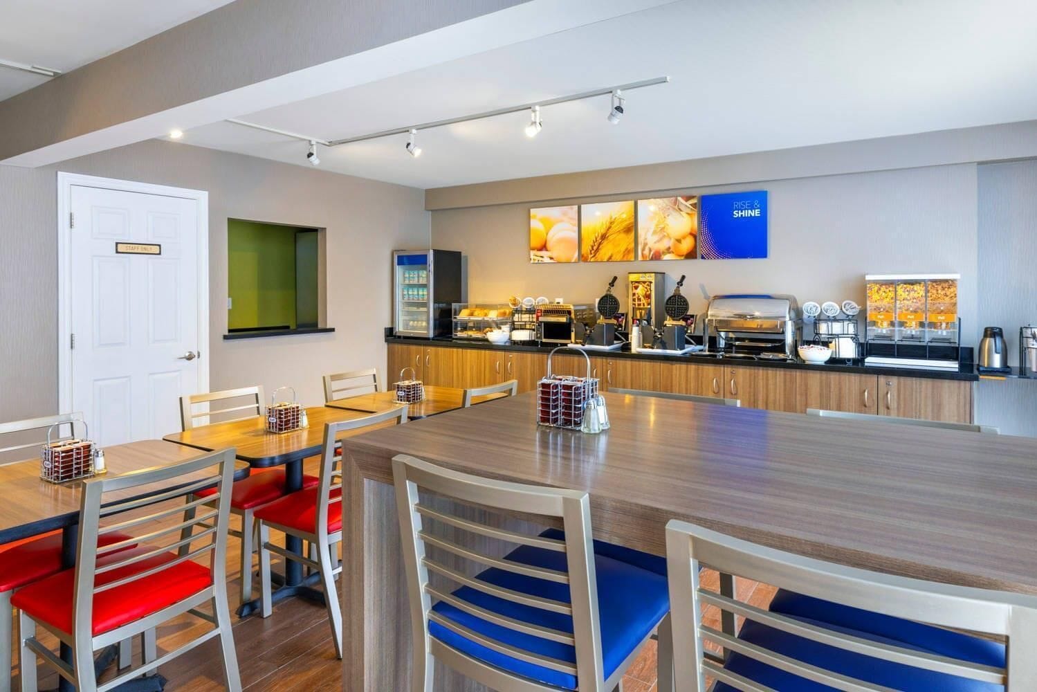 A modern hotel room with a dining area featuring a long wooden table and chairs with red and blue cushions. A kitchenette with a coffee machine and a TV mounted on the wall is visible.