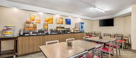 A modern dining area with wooden tables and red cushioned chairs, a kitchen counter with various appliances, and wall-mounted artwork.