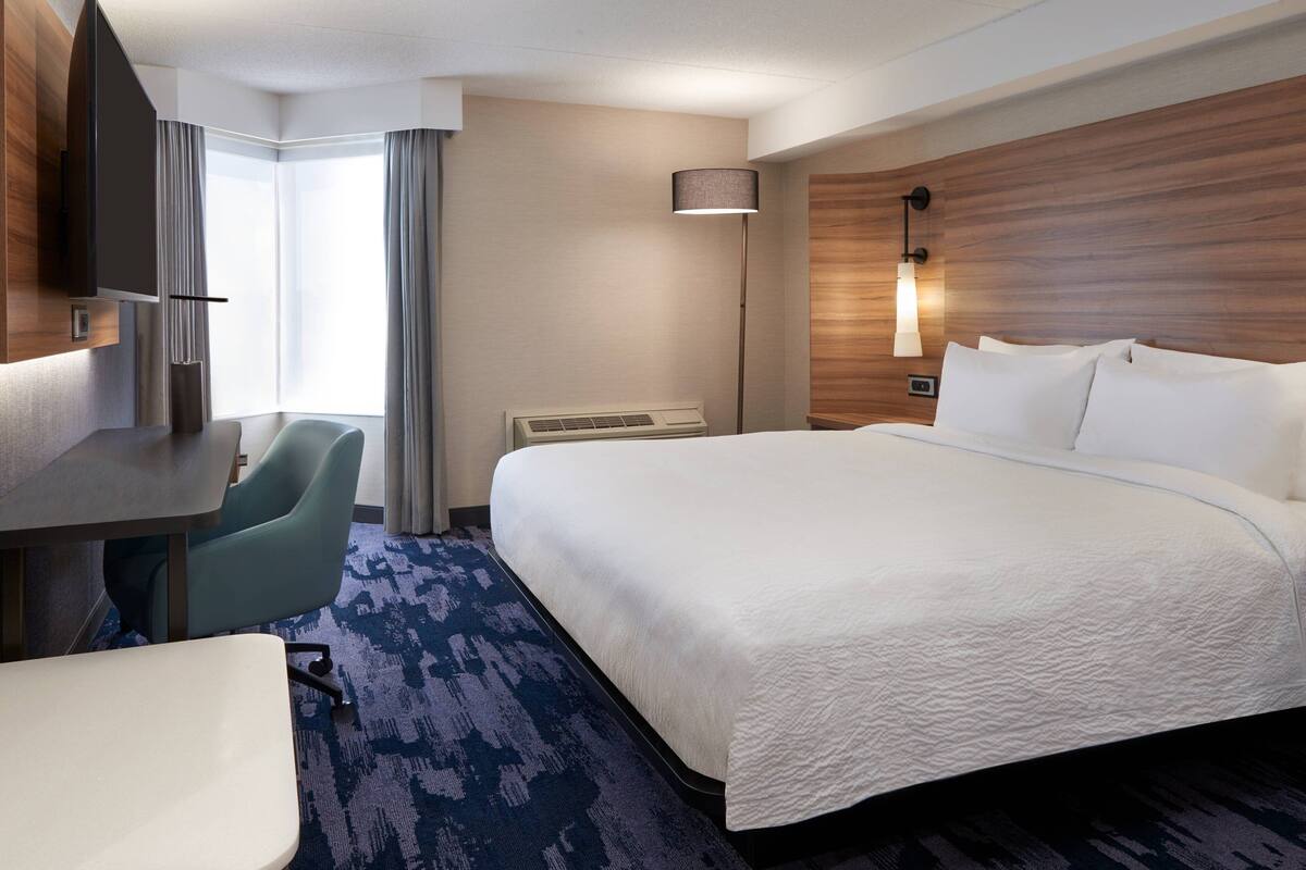 A hotel room with a large bed, a desk, a chair, a TV, and a lamp.