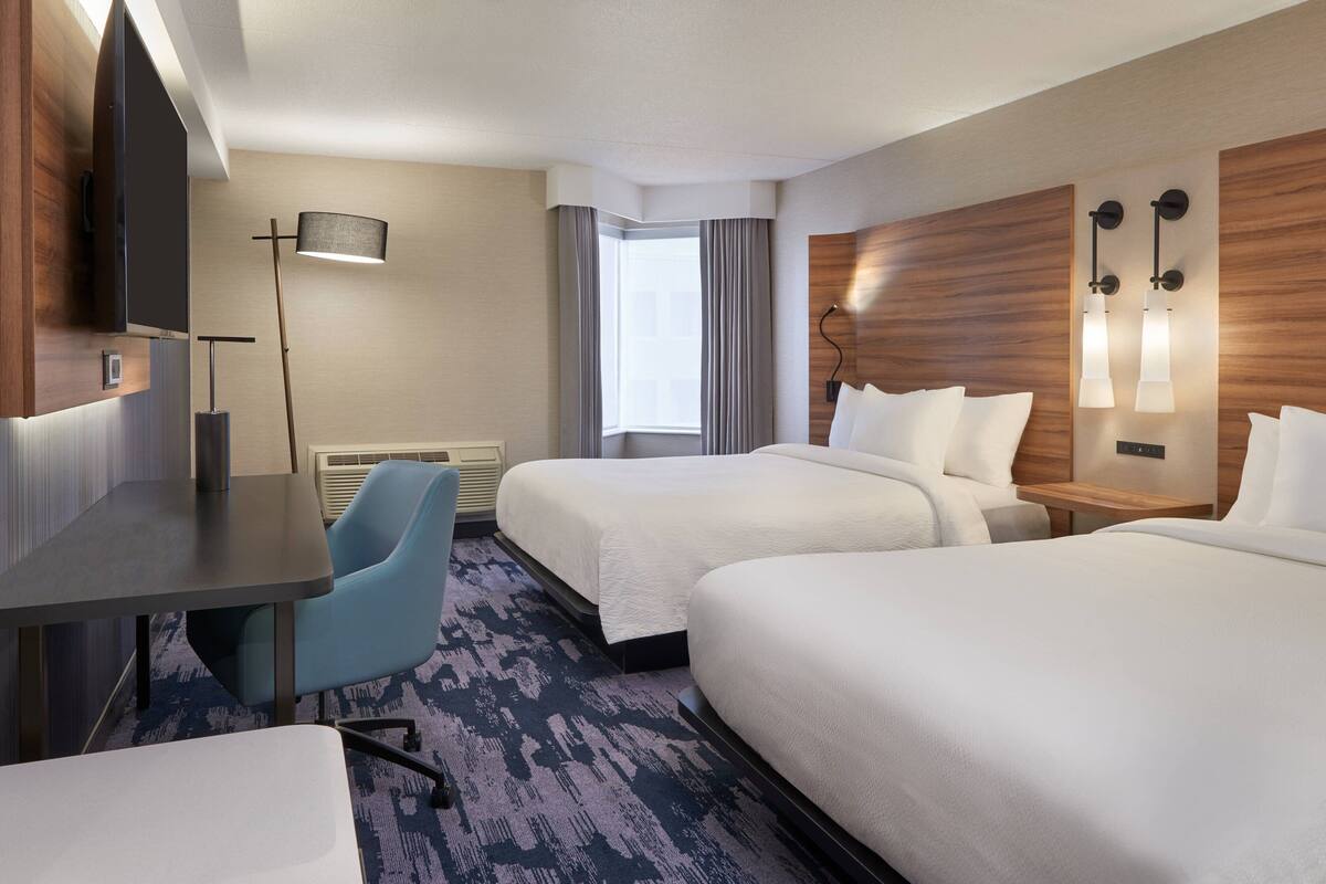 A hotel room with two beds, a desk, a chair, a flat-screen TV, a lamp, and a window with curtains.