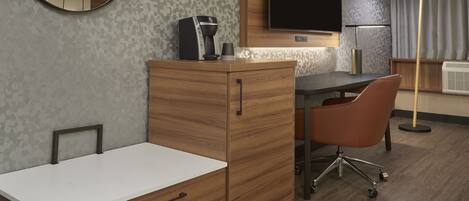 A modern hotel room with a wooden desk, a chair, a flat-screen TV, a coffee machine, and a bedside table.