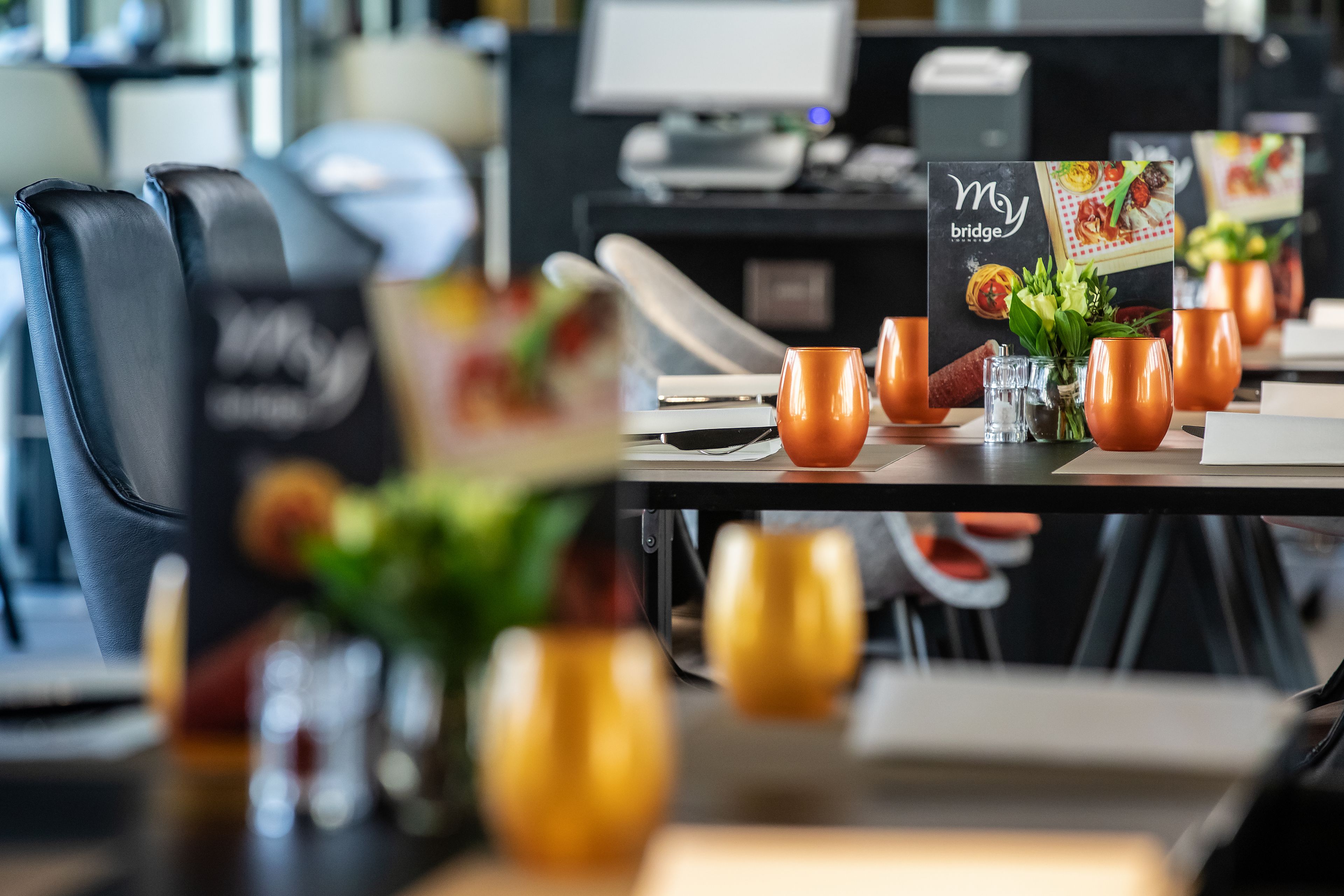 Une scène de restaurant avec un présentoir à menus, des tasses oranges et un ordinateur portable posé sur la table.