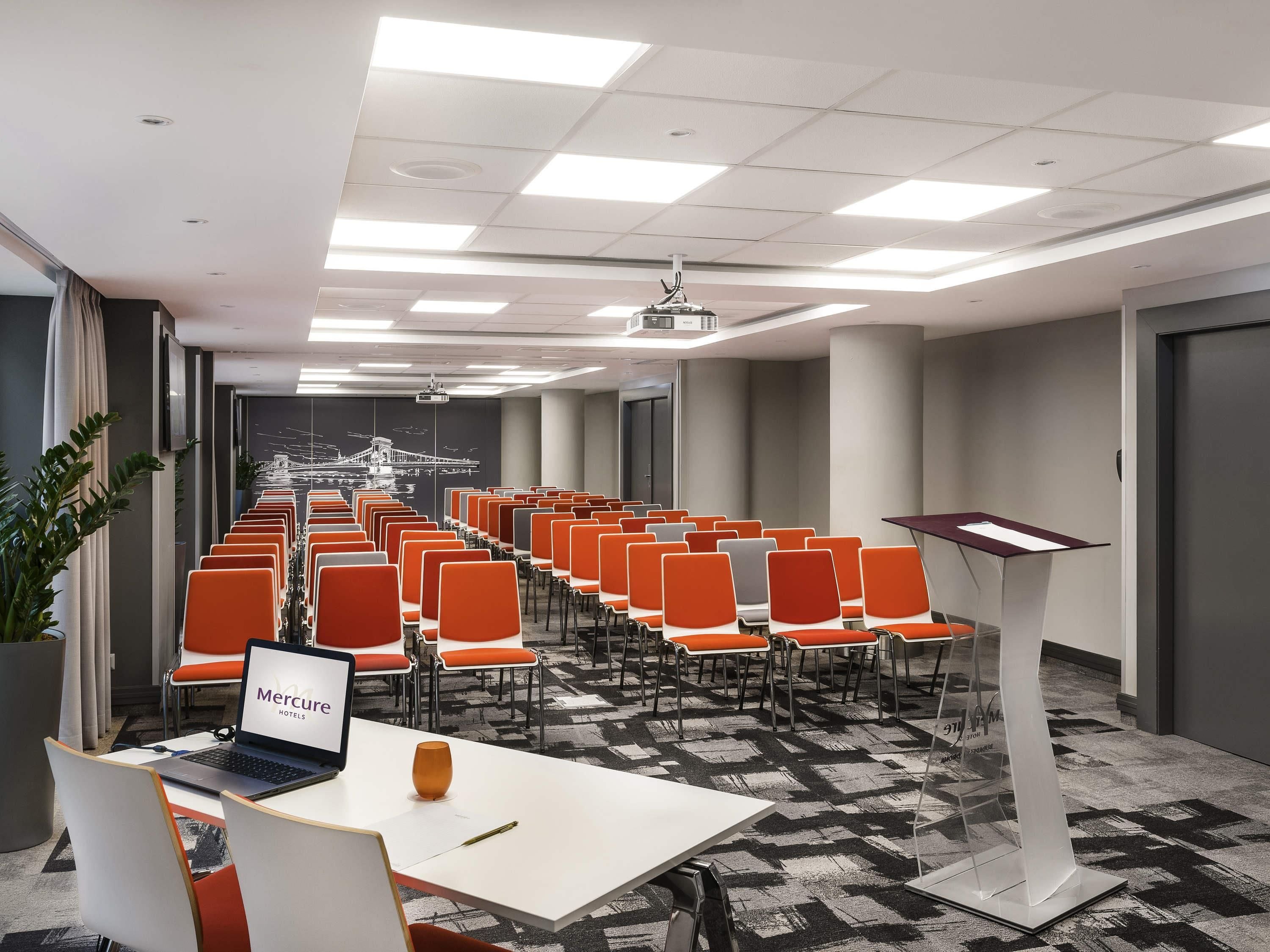Une salle de conférence avec des rangées de chaises orange, une table blanche sur laquelle se trouve un ordinateur portable et un pupitre.